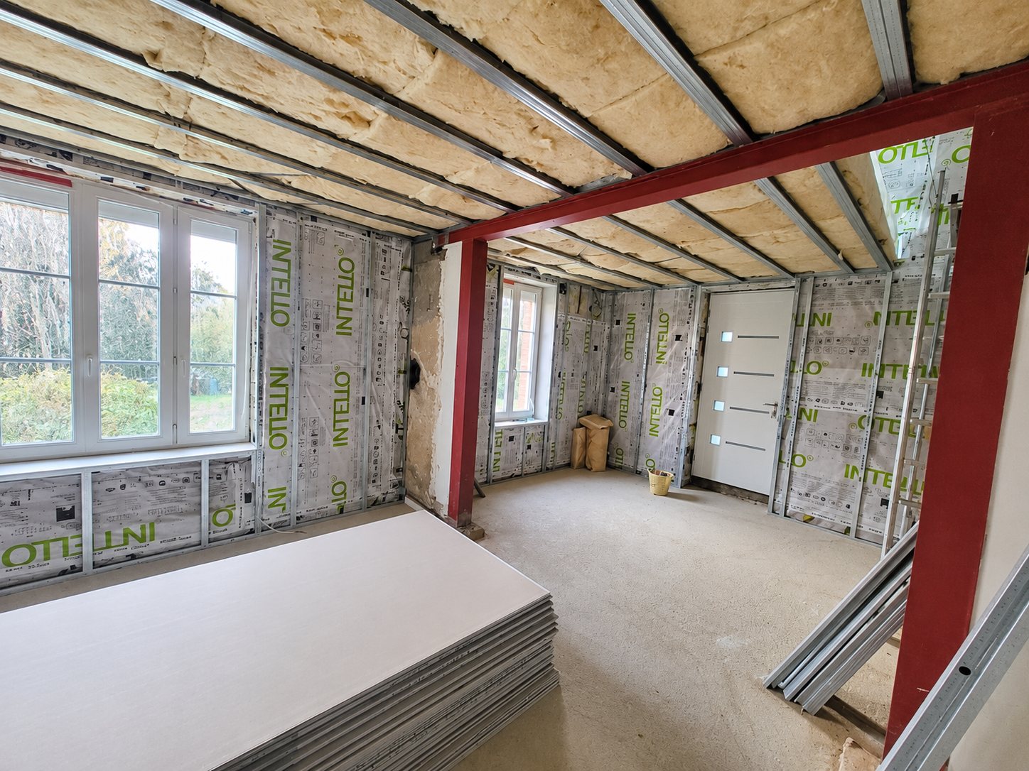Pièce en cours de rénovation avec membrane d'isolation Intello sur les murs, laine en plafond, poutre rouge et pile de plaques de plâtre au premier plan.