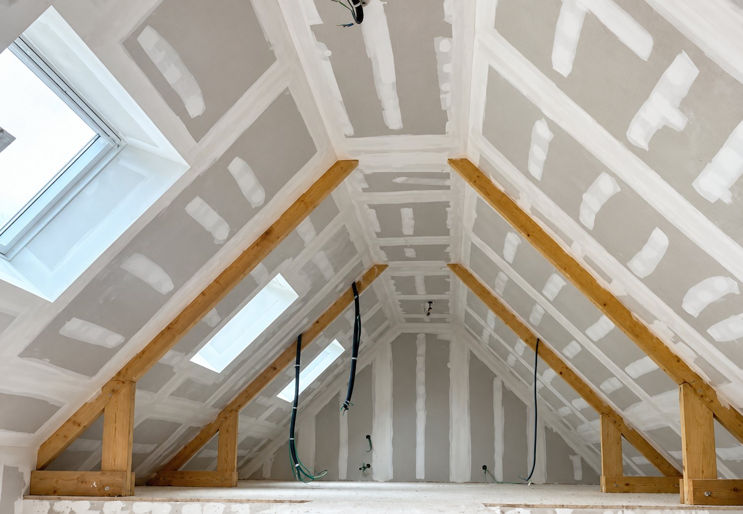 Intérieur de combles en cours d'aménagement, avec plaques de plâtre jointoyées, poutres en bois apparentes et plusieurs fenêtres de toit.