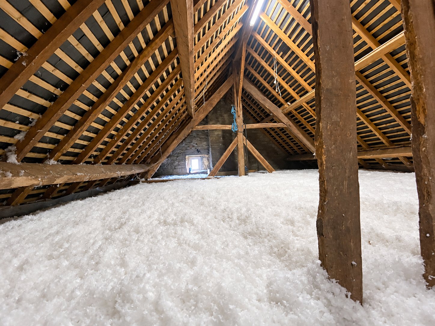 Vue de combles sous une charpente en bois dont le sol est recouvert d'une épaisse couche d'isolant soufflé blanc.