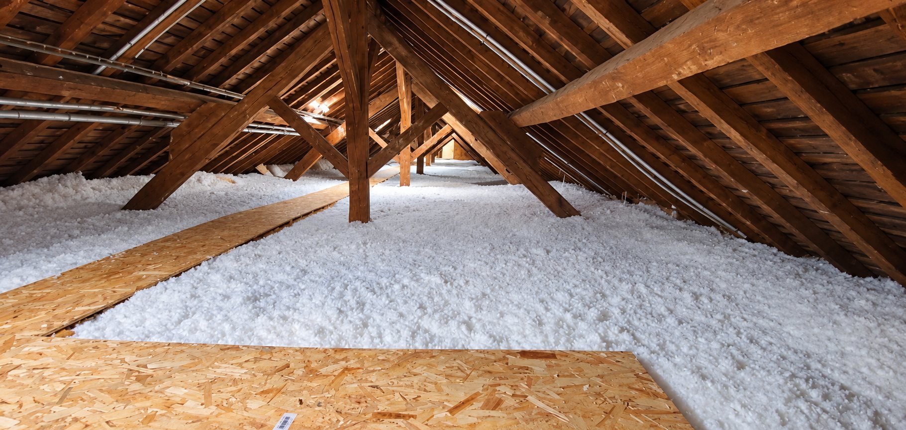 Vue de combles sous une charpente en bois, dont le sol est recouvert d'une épaisse couche d'isolant soufflé blanc et de panneaux de circulation en OSB.