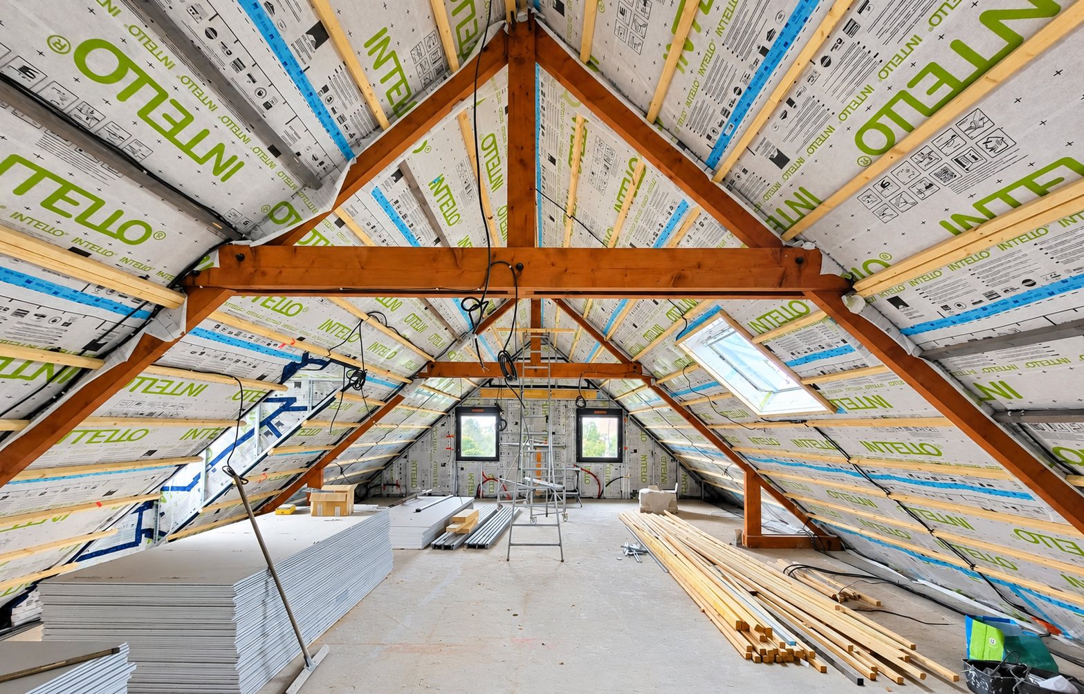 Vaste comble en cours d'aménagement dont les rampants sont couverts d'une membrane Intello, avec charpente en bois, échafaudage, plaques de plâtre et lattes au sol.