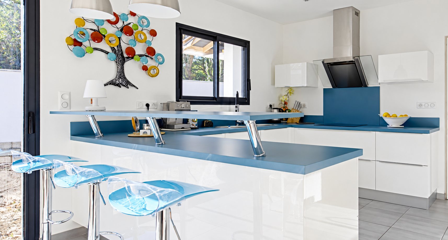 Cuisine ouverte aux meubles blancs laqués et au plan de travail bleu, avec un bar et trois tabourets bleu transparent, sous un arbre décoratif coloré au mur.