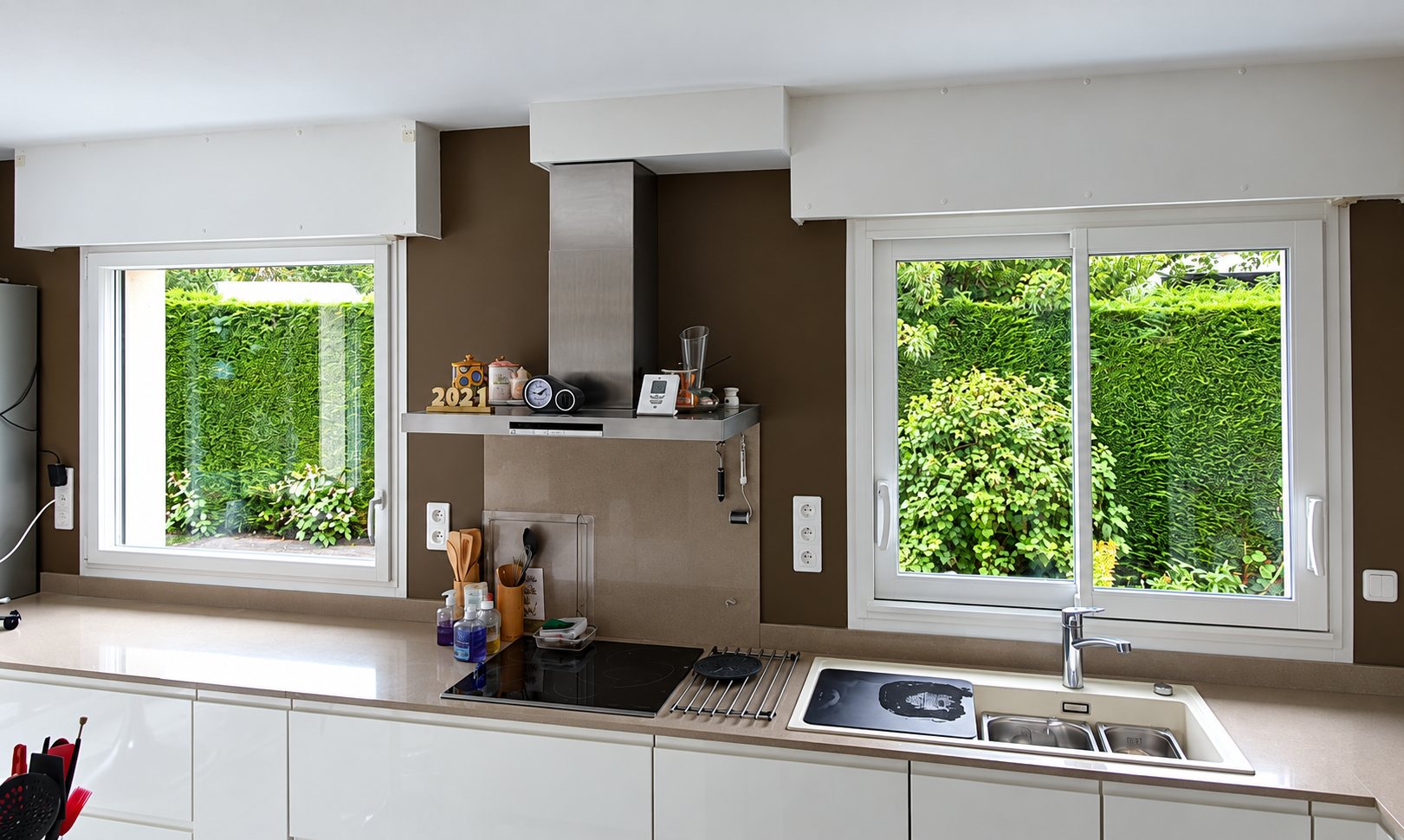 Une cuisine moderne aux murs bruns et aux meubles blancs, équipée de deux fenêtres en PVC blanc qui donnent sur une haie verte.