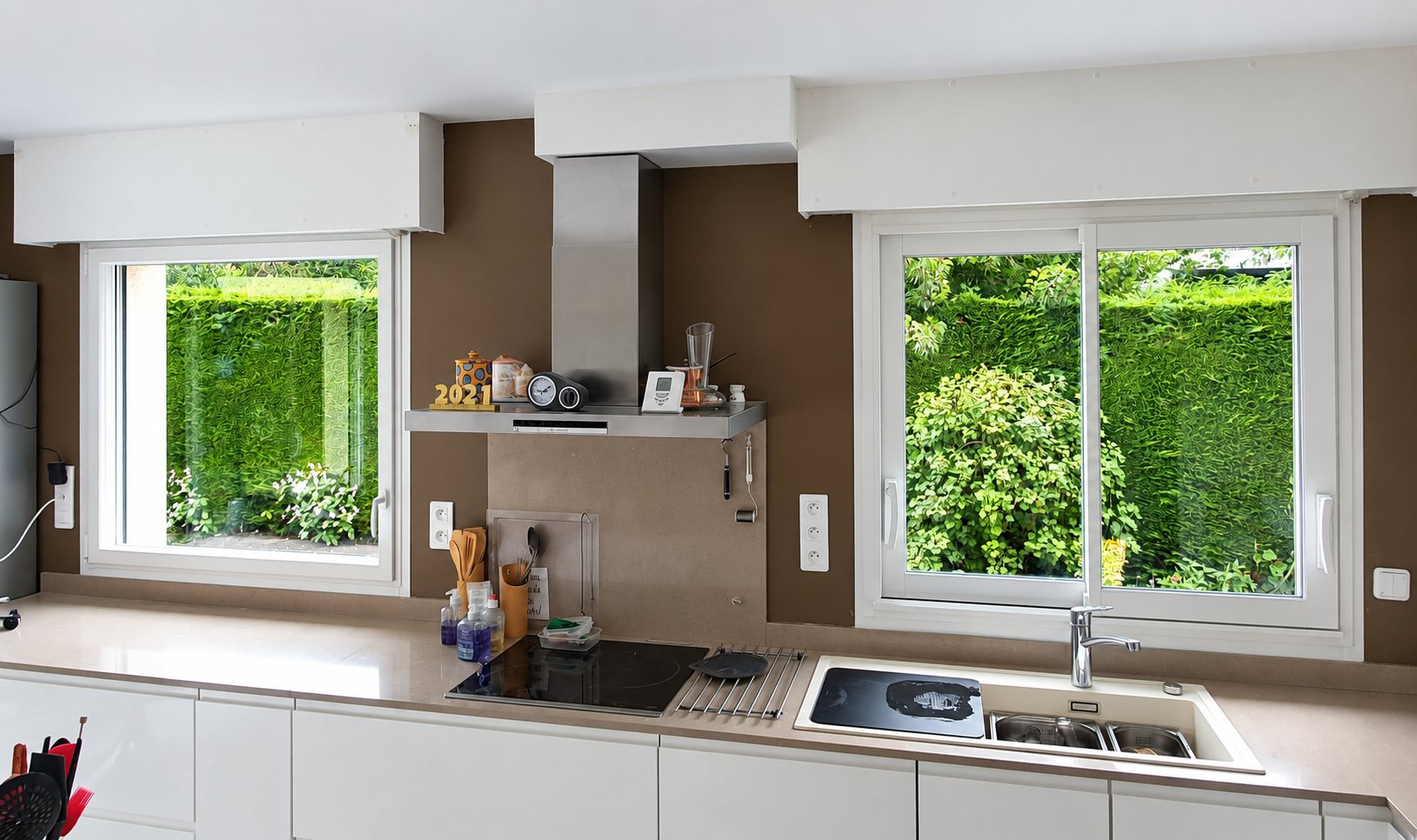 Une cuisine moderne aux murs bruns et aux meubles blancs équipée de deux fenêtres en PVC blanc qui donnent sur une haie verte et un massif fleuri.