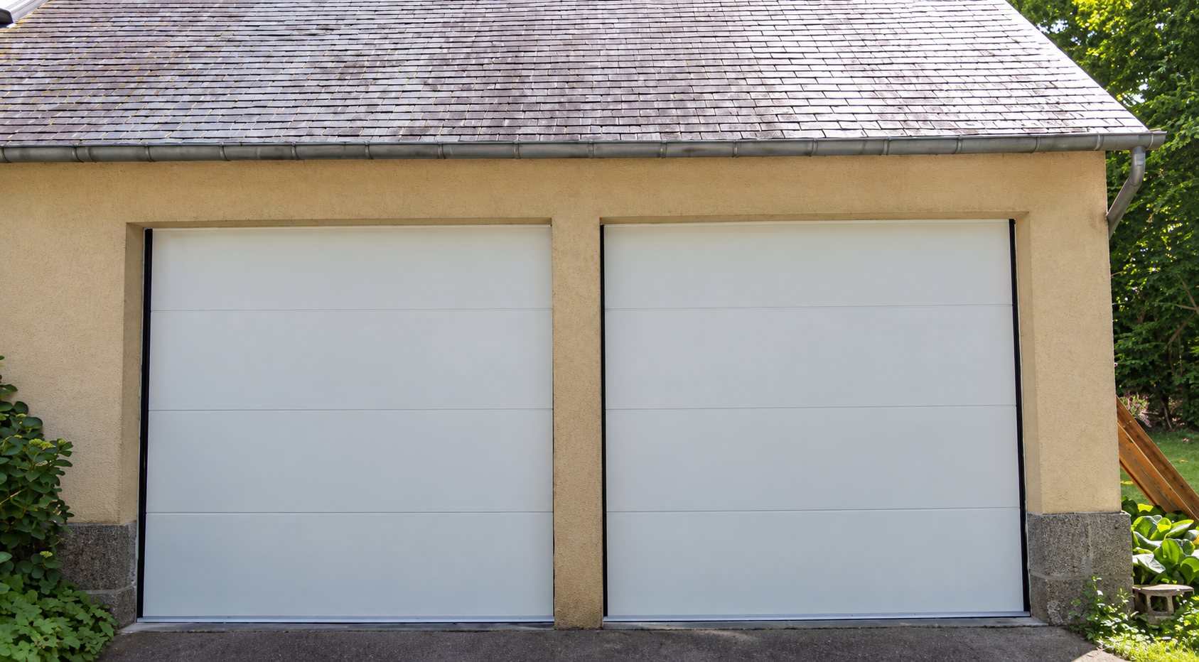 Deux portes de garage sectionnelles blanches posées dans une façade en enduit beige sous un toit en ardoise.
