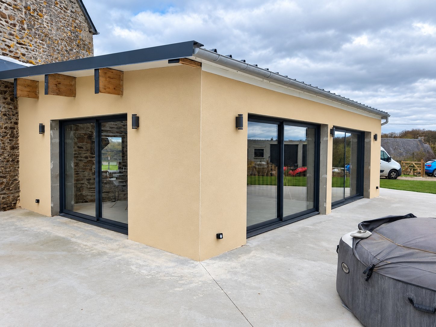 Extension récente à l'enduit beige accolée à une maison en pierre, avec de grandes baies vitrées coulissantes anthracite donnant sur une terrasse en béton.