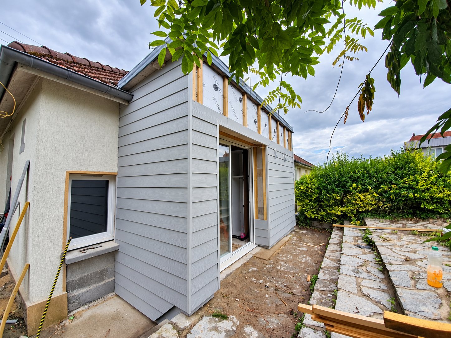 Chantier d'extension d'une petite maison dont la façade est en partie recouverte d'un bardage gris à lames horizontales, avec une porte-fenêtre et un jardin en pierre au premier plan.