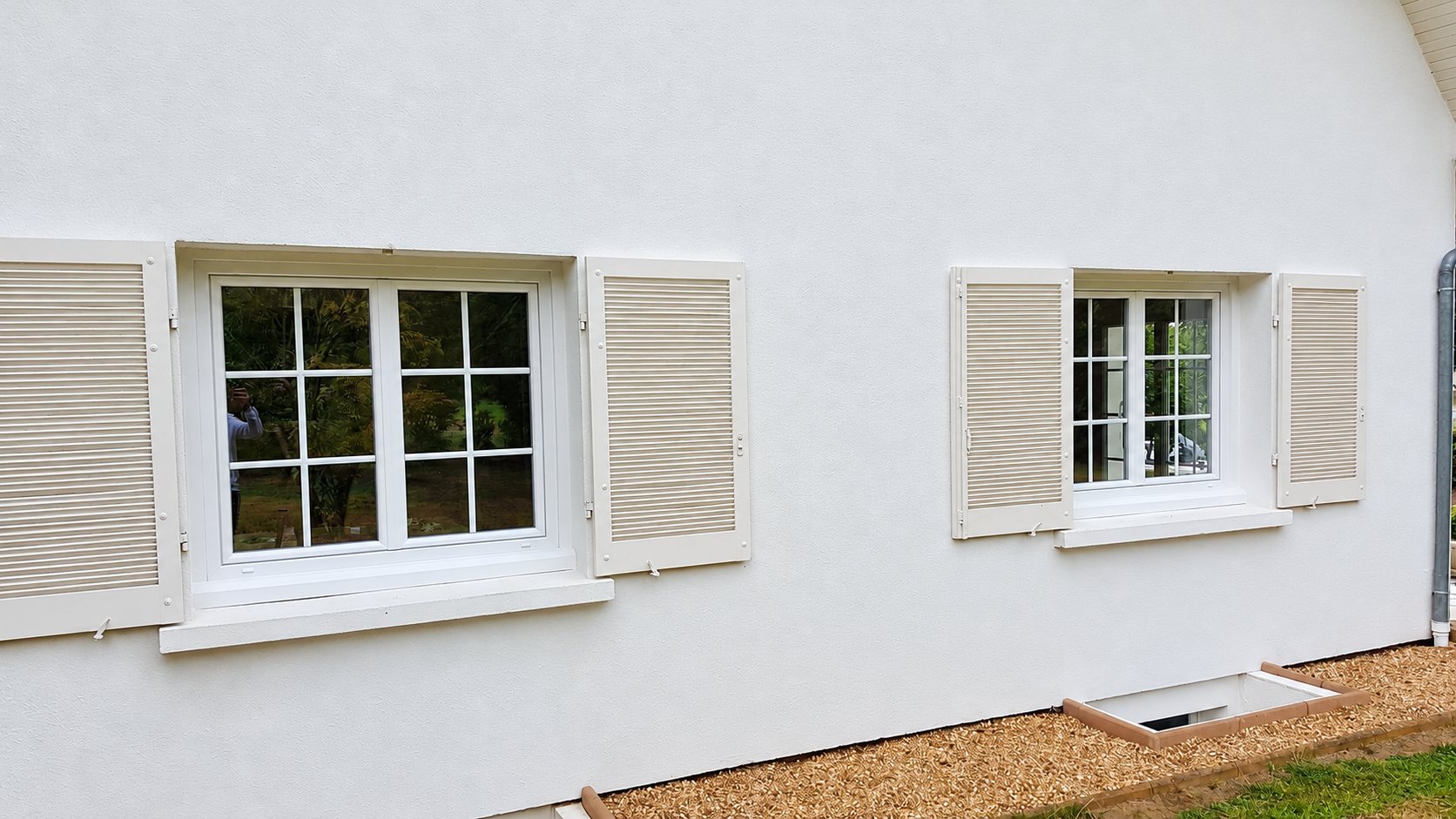 Façade d'une maison enduite de blanc avec deux fenêtres à petits carreaux et des volets persiennes beiges ouverts, au-dessus d'un sol de gravier.