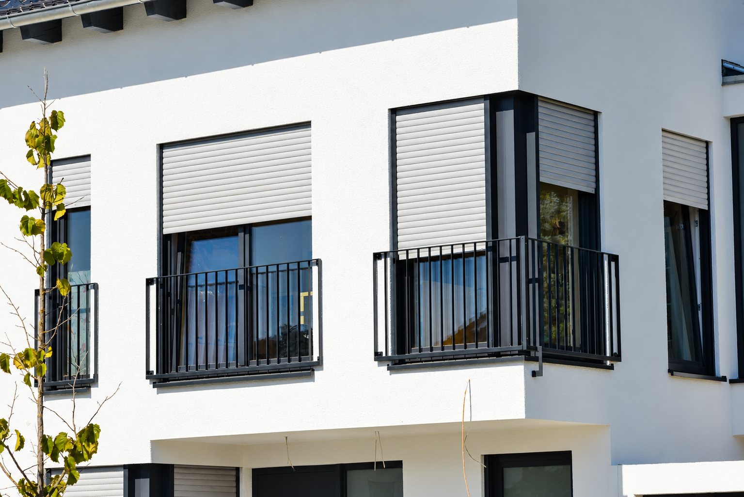 Façade d'un immeuble moderne blanc équipé de volets roulants beiges et de garde-corps métalliques anthracite devant les fenêtres.
