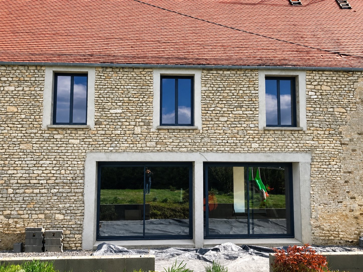 Façade d'une maison en pierre apparente et toit de tuiles rouges, équipée de trois fenêtres et de deux grandes baies vitrées coulissantes noires.