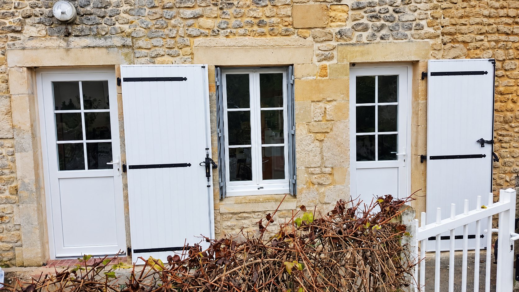 Façade en pierre apparente équipée de deux portes vitrées blanches, d'une fenêtre à petits carreaux et de volets blancs avec ferrures noires.
