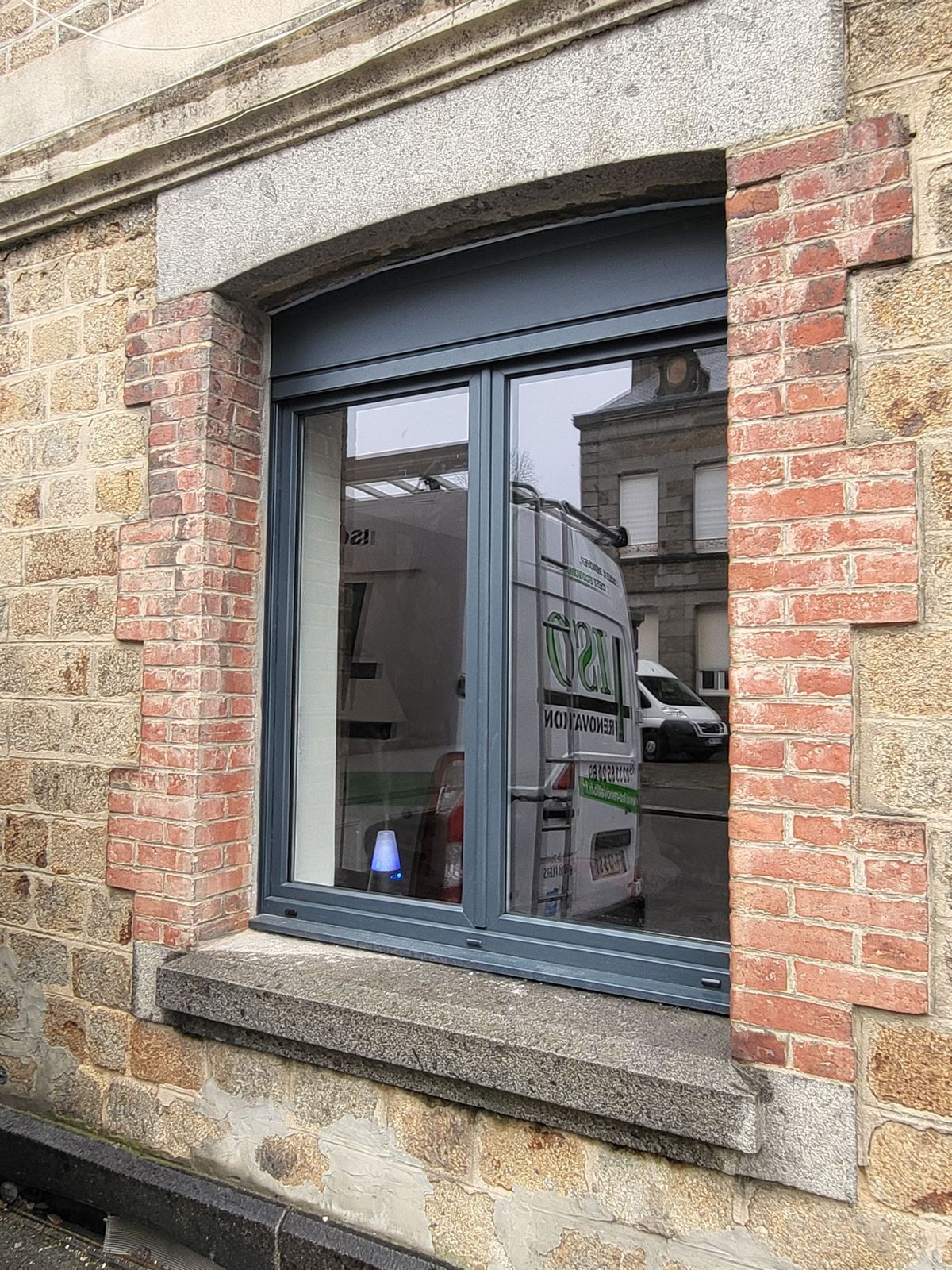 Fenêtre grise à deux vantaux avec volet roulant, installée dans une façade en pierre et briques rouges, où se reflète une camionnette de l'entreprise.