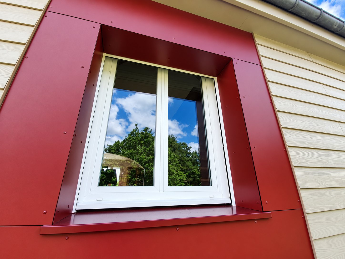 Fenêtre blanche à deux vantaux entourée d'un habillage métallique rouge bordeaux, sur une façade en clin beige, avec reflet du ciel et des arbres dans la vitre.