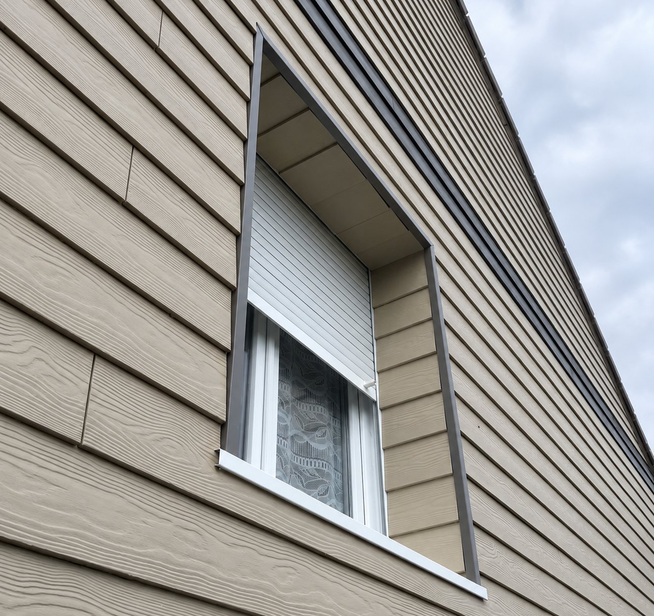 Vue en contre-plongée d'une façade habillée de bardage gris-beige à clin, avec une fenêtre blanche équipée d'un volet roulant partiellement baissé.