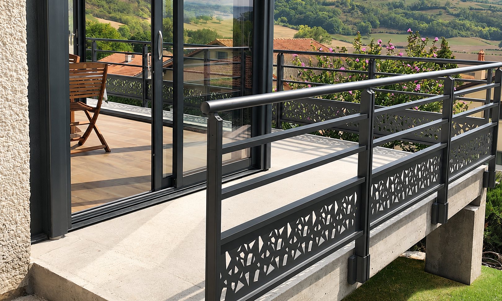 Un garde-corps gris anthracite aux panneaux ajourés borde une terrasse en béton devant une grande baie vitrée coulissante, avec vue sur la campagne.
