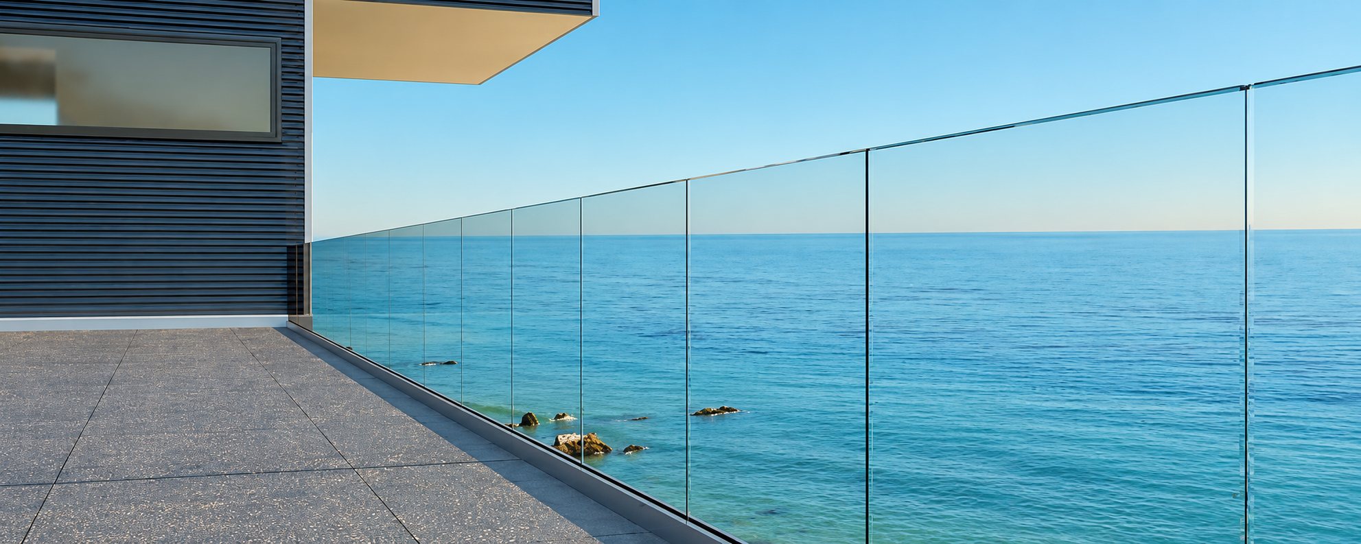 Une terrasse dallée d'une maison moderne est bordée d'un garde-corps en verre transparent qui donne sur la mer calme.