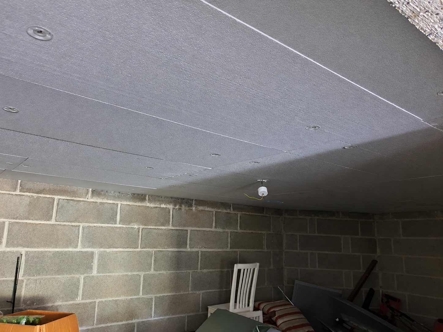 Plafond d'une cave recouvert de panneaux isolants gris, au-dessus de murs en parpaings, avec une chaise blanche et un carton au sol.