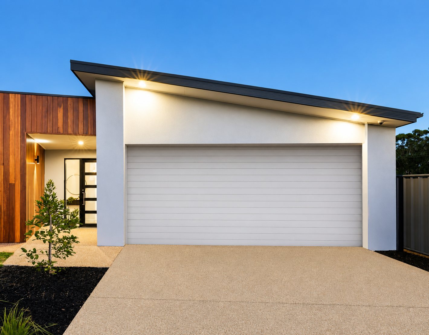 Façade d'une maison contemporaine au crépuscule avec une porte de garage blanche à lames horizontales, un bardage en bois et une allée en béton beige.