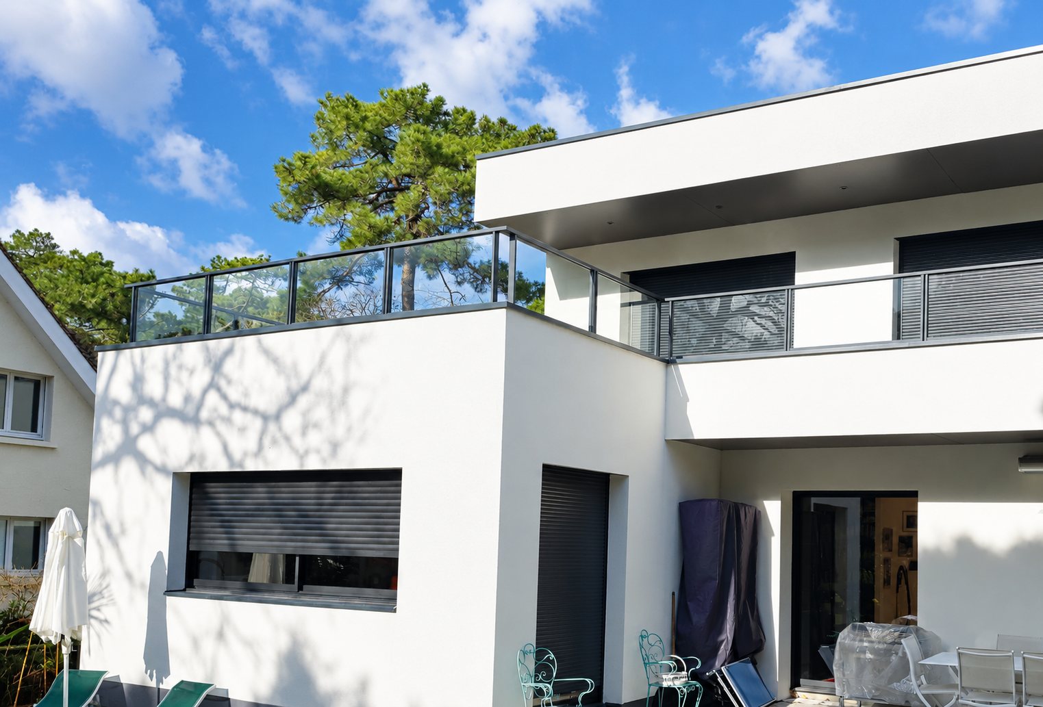 Façade d'une maison contemporaine blanche à toit plat, équipée de garde-corps en verre et en aluminium et de volets roulants gris anthracite, avec un pin en arrière-plan.