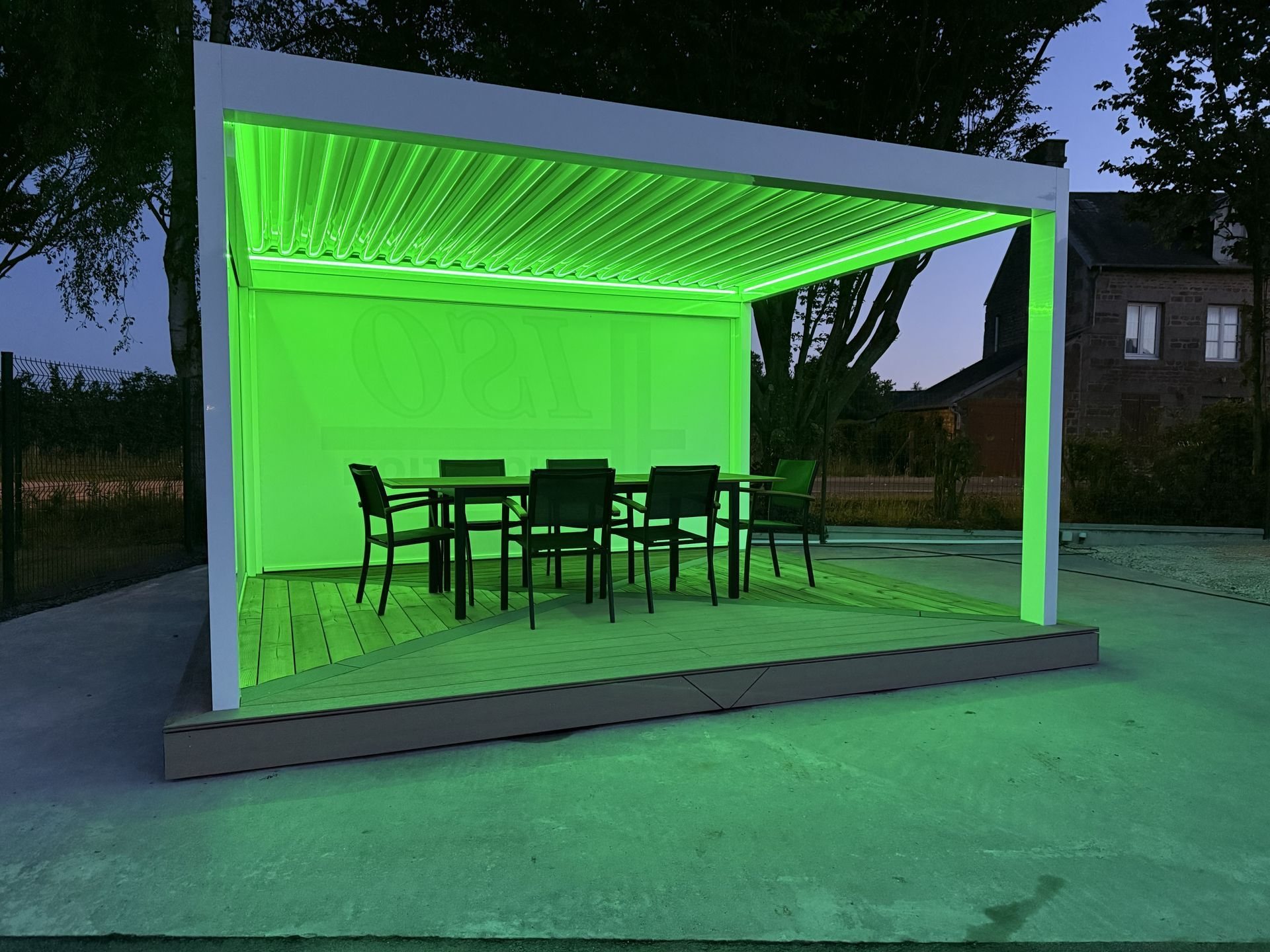 Pergola bioclimatique blanche à lames orientables, éclairée en vert par des LED, abritant une table et six chaises sur une terrasse en bois au crépuscule.