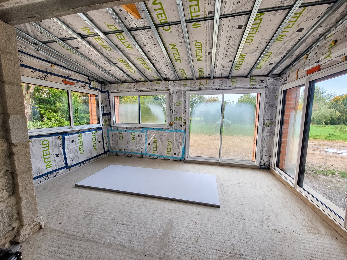 Intérieur d'une pièce en chantier dont les murs et le plafond sont isolés avec une membrane Intello, avec des fenêtres coulissantes et une plaque de plâtre posée au sol.