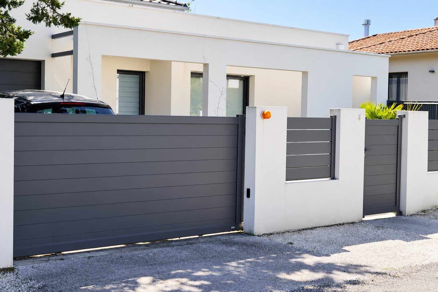 Un portail coulissant et un portillon en aluminium gris à lames horizontales, encadrés de piliers blancs, devant une maison contemporaine.