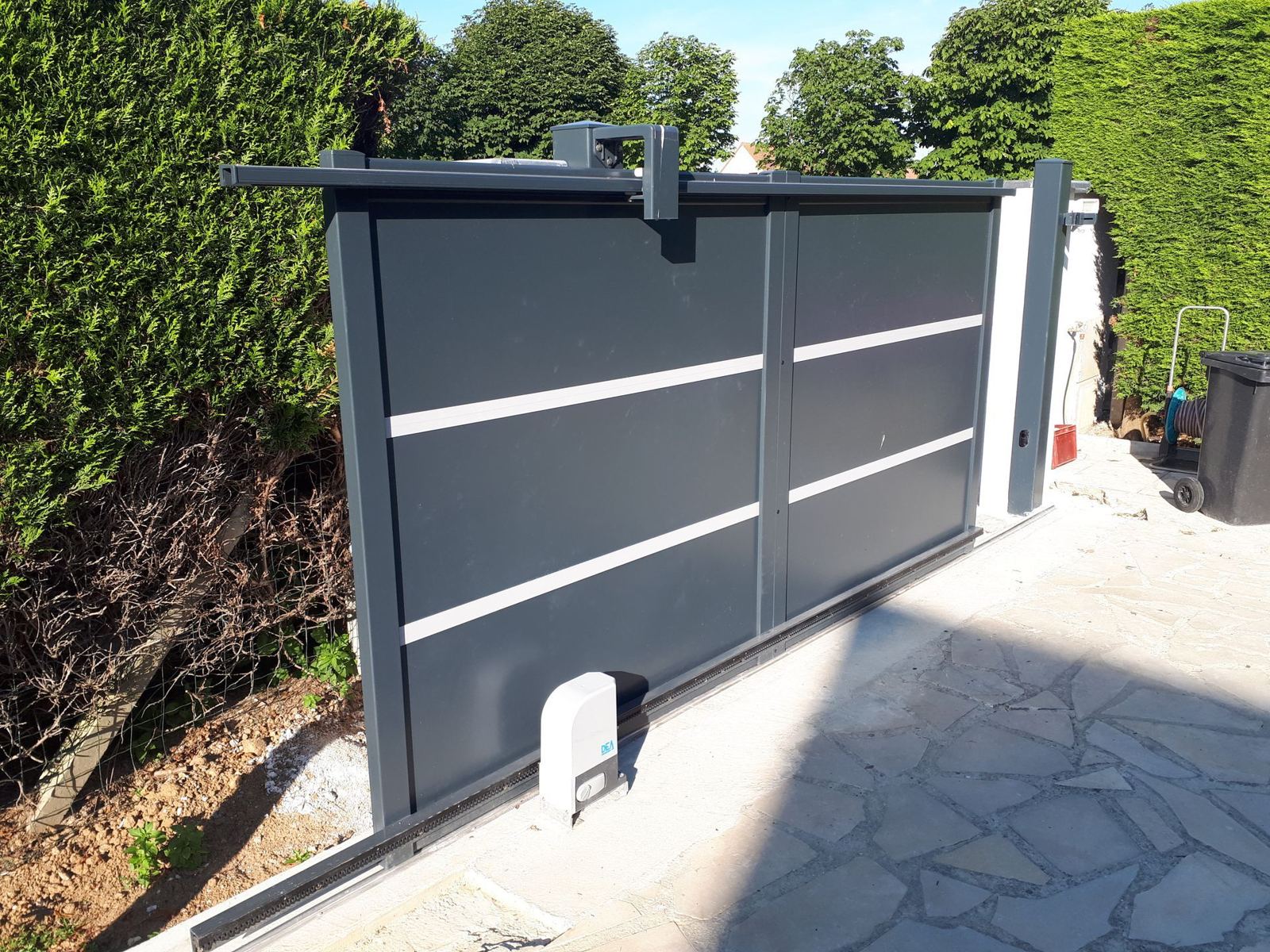 Un portail coulissant en aluminium gris anthracite avec lignes claires et moteur au sol, posé entre deux haies, devant une allée en pierres.