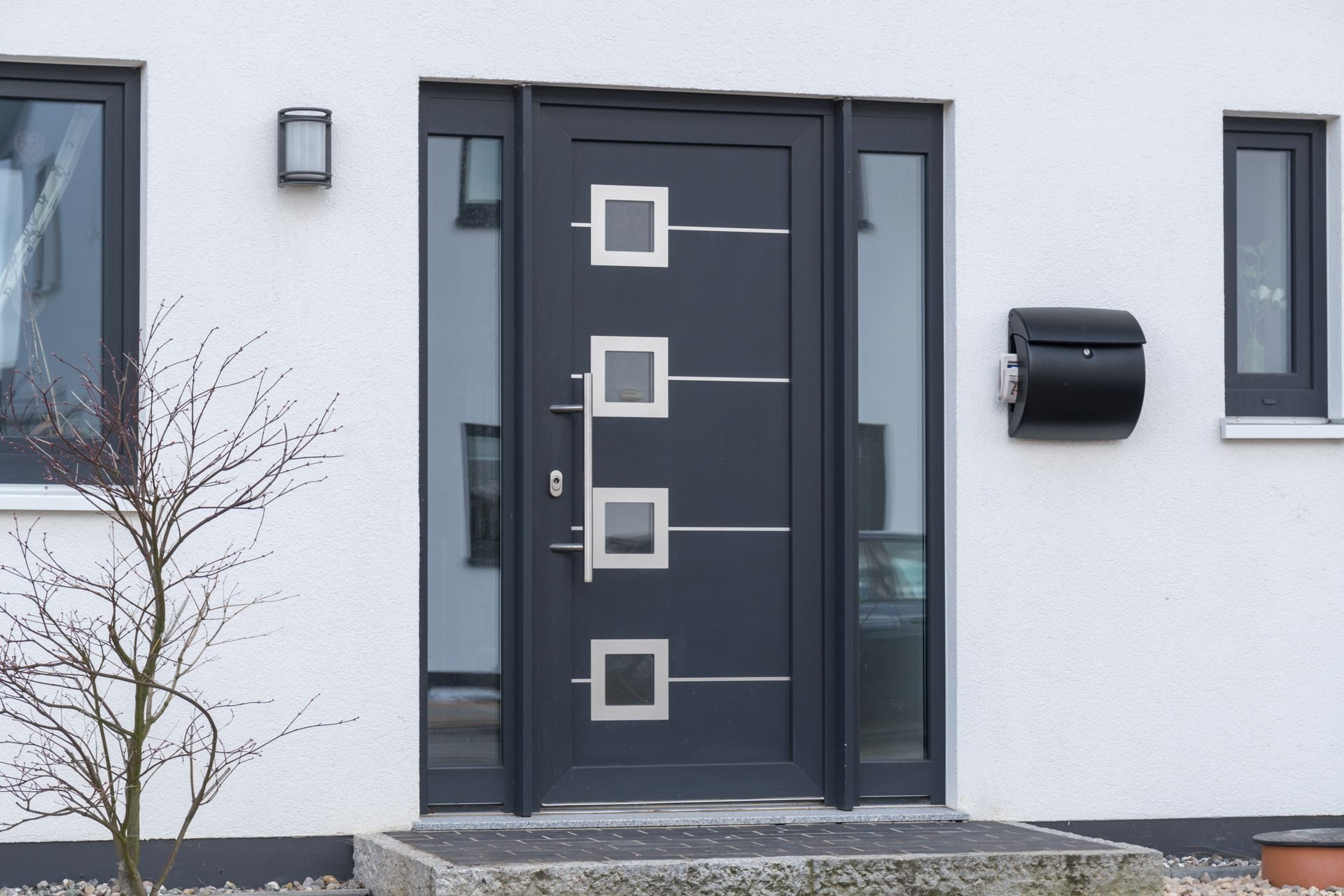 Porte d'entrée gris anthracite à quatre petits vitrages carrés encadrés d'inox, bordée de deux panneaux vitrés, sur une façade blanche avec boîte aux lettres noire.