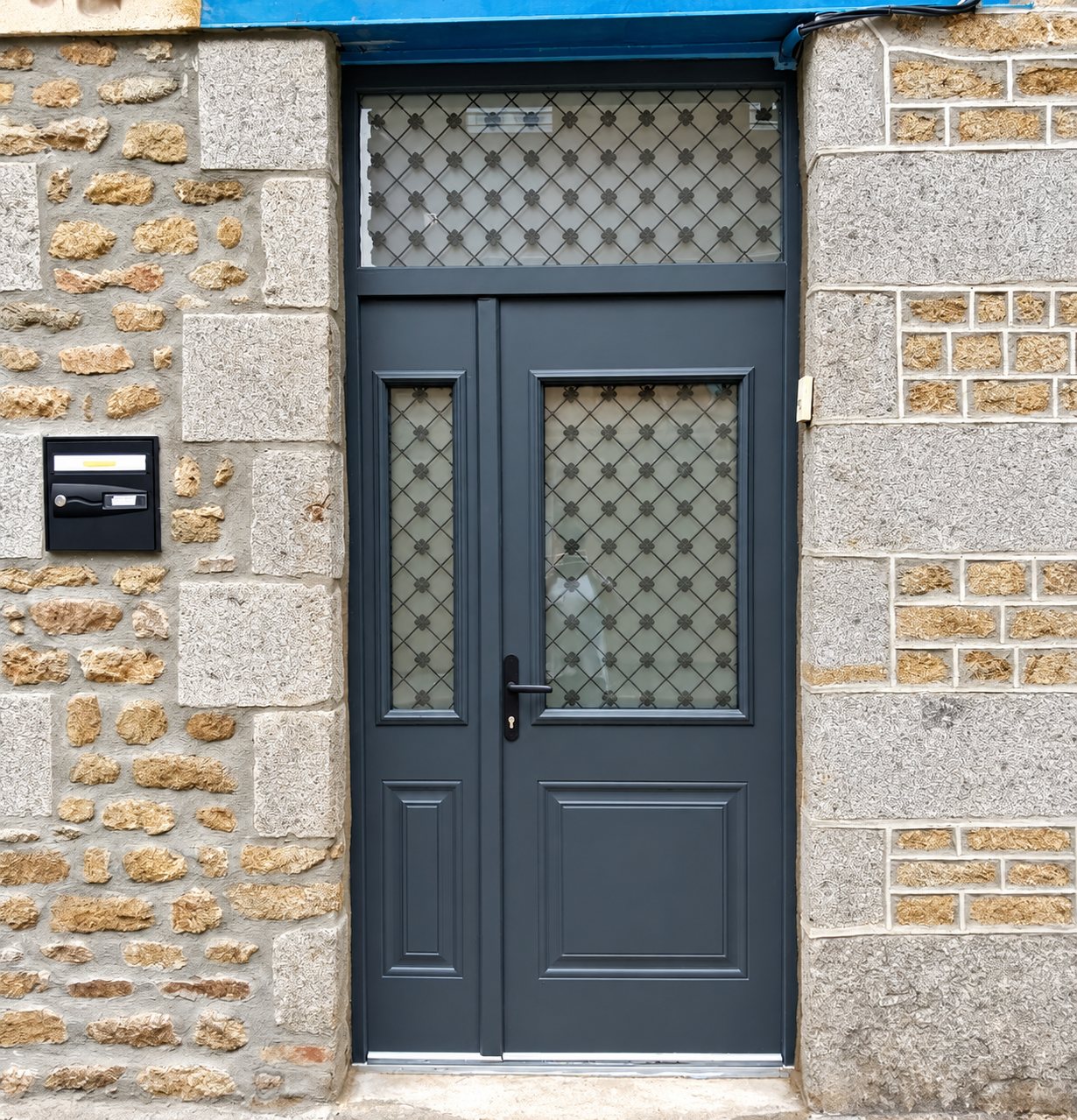 Porte d'entrée gris anthracite à deux vantaux avec vitrages décoratifs et imposte, encadrée de murs en pierre de granit, avec une boîte aux lettres noire à gauche.