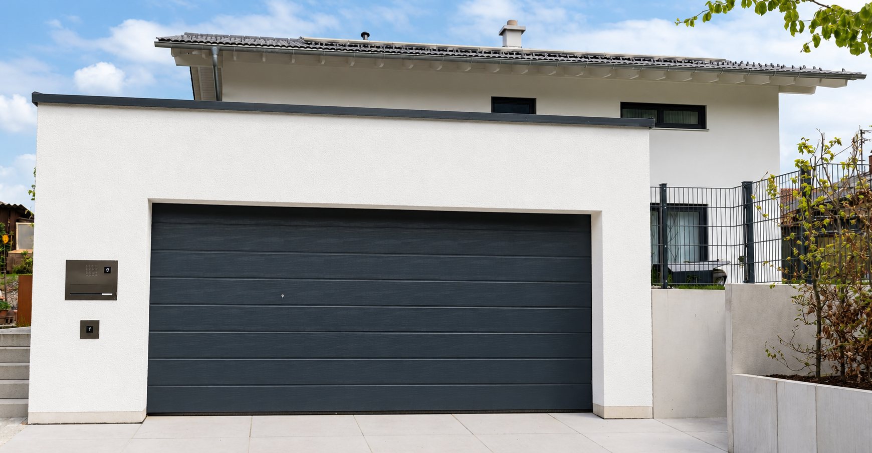 Une porte de garage sectionnelle gris anthracite installée dans un garage à façade blanche, devant une maison moderne avec clôture métallique.