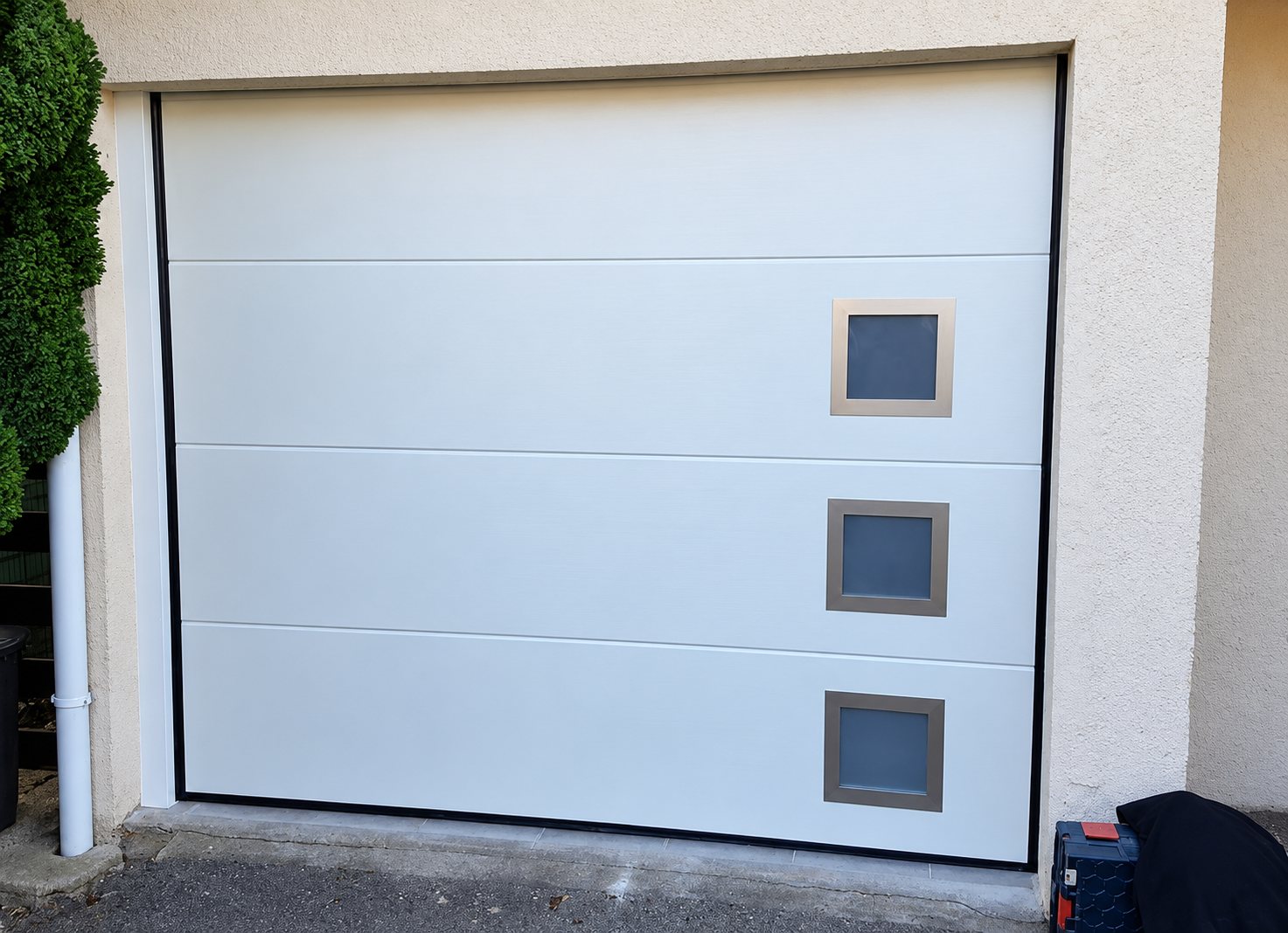 Porte de garage sectionnelle blanche installée dans une façade crépie, équipée de trois petits hublots carrés à cadre métallique.