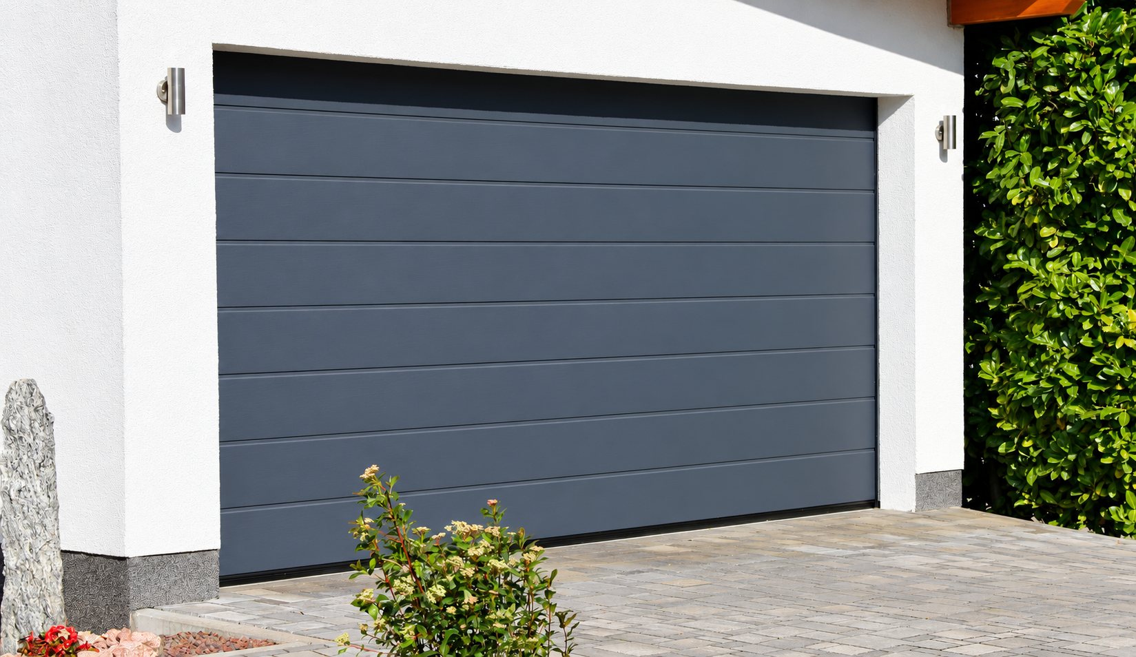 Une porte de garage sectionnelle gris anthracite installée dans une façade blanche, devant une allée pavée et une haie verte.