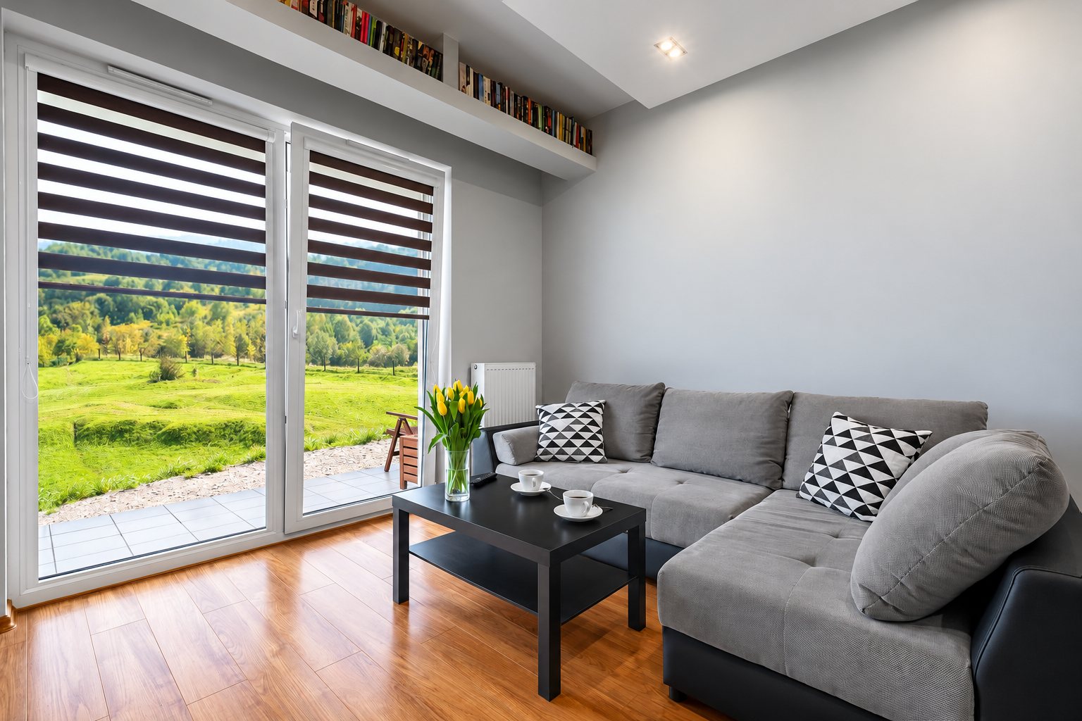Un salon lumineux avec canapé d'angle gris, table basse noire et bouquet de tulipes jaunes, dont la baie vitrée équipée de stores jour-nuit donne sur une prairie verte.