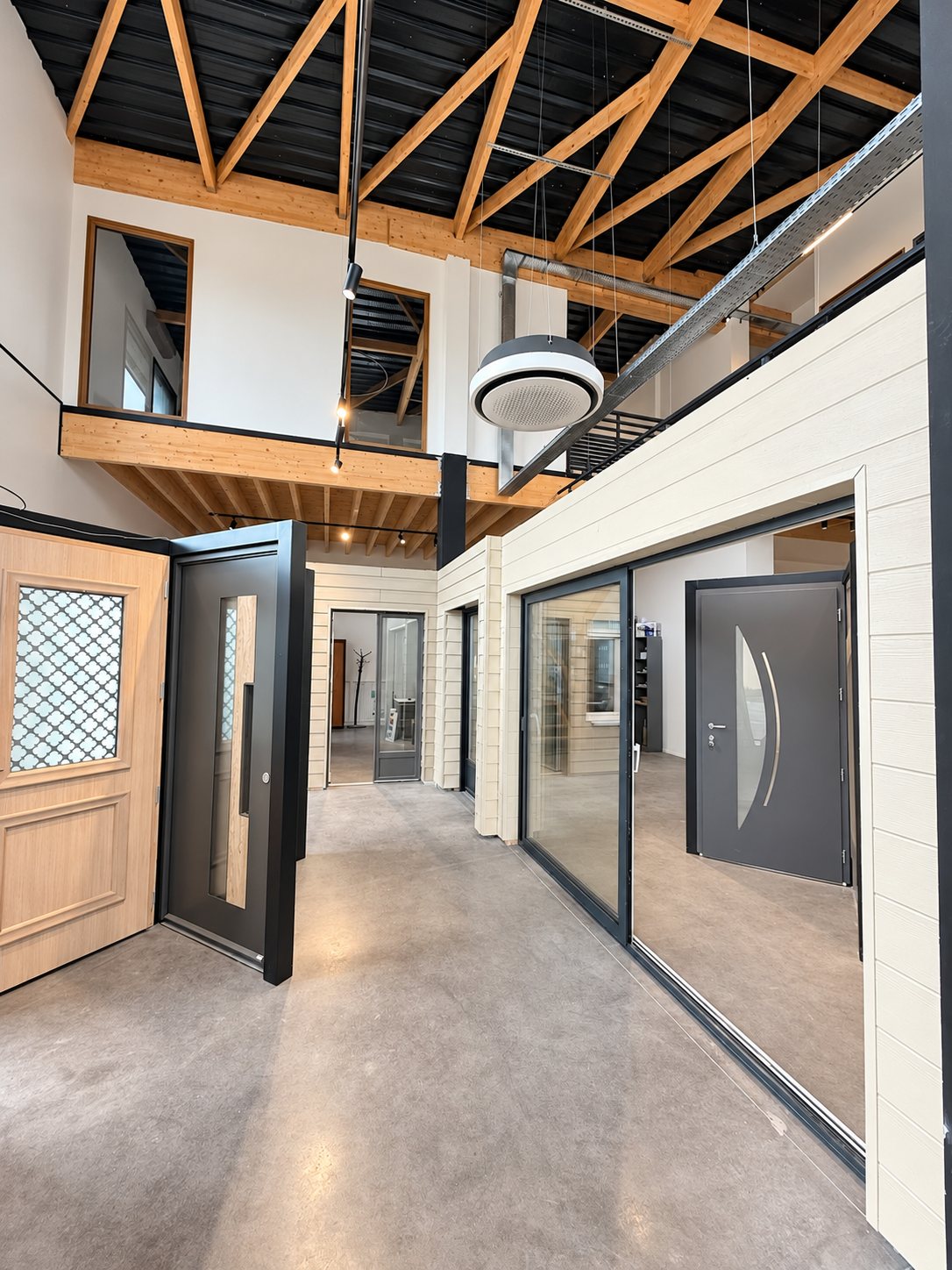 Intérieur d'un showroom lumineux avec portes d'entrée en bois et en aluminium gris, baie coulissante et mezzanine sous une charpente apparente.