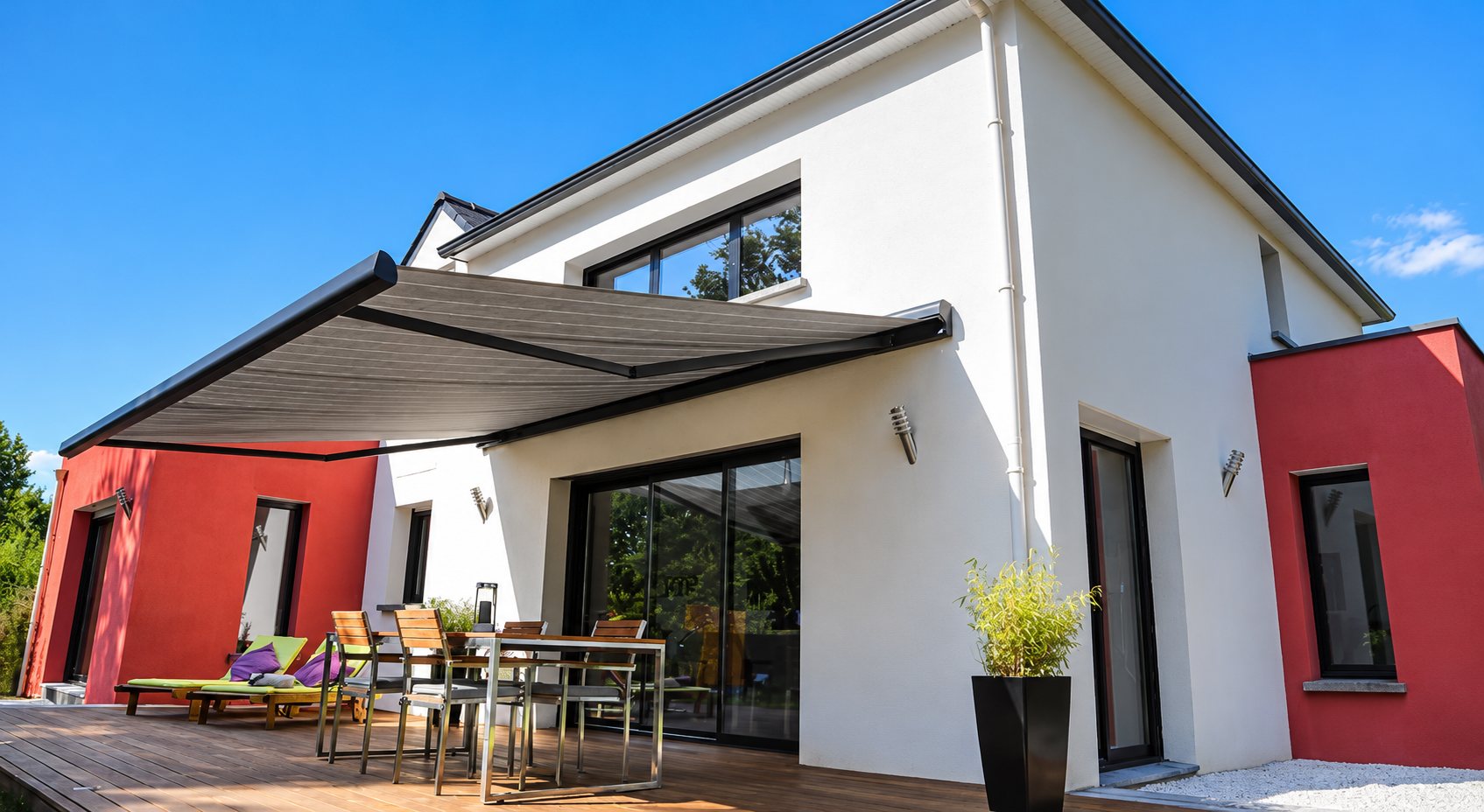 Un grand store banc gris anthracite est déployé au-dessus d'une terrasse en bois avec table et chaises, devant une maison moderne blanche et rouge sous un ciel bleu.