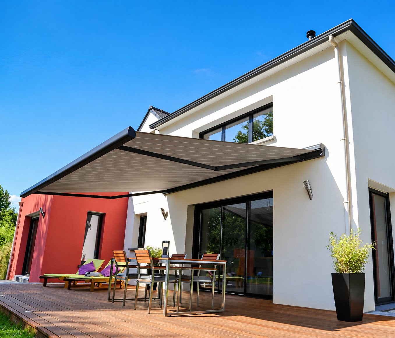 Un grand store banne gris déployé protège une terrasse en bois équipée d'une table, de chaises et de transats devant une maison blanche et rouge.