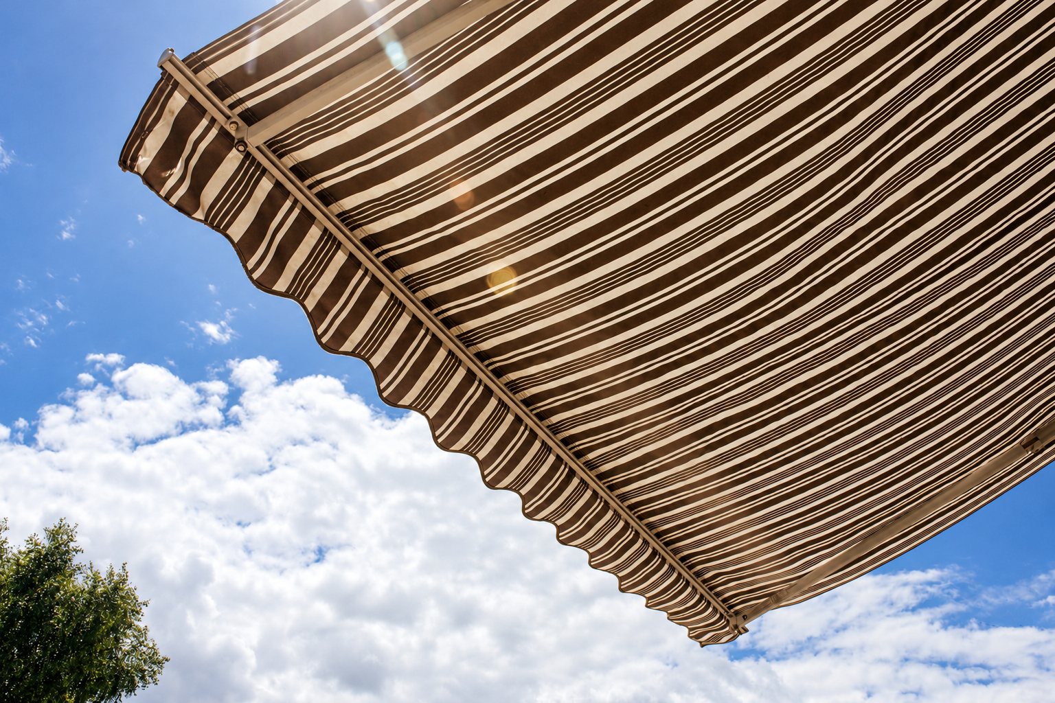 Vue en contre-plongée d'un store banne à rayures marron et beige déployé, avec un ciel bleu parsemé de nuages et la cime d'un arbre en arrière-plan.