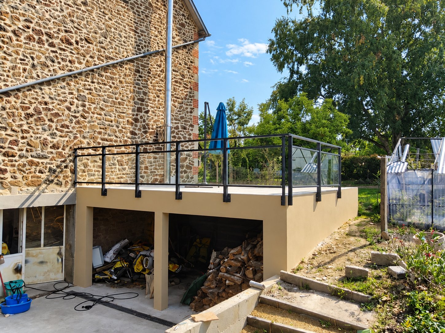 Une terrasse surélevée aux murs enduits beige, dotée d'un garde-corps en verre et métal noir, est construite contre une maison en pierre, avec du bois de chauffage stocké en dessous.