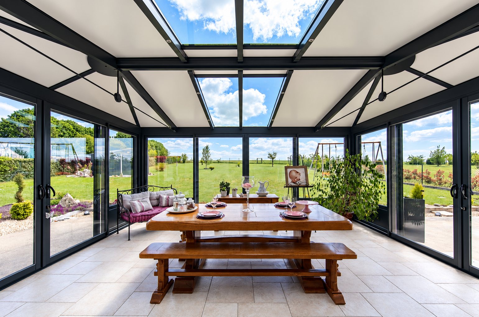 Intérieur lumineux d'une véranda à structure sombre et toit vitré, meublée d'une grande table en bois avec bancs et d'un banc à coussins, ouverte sur un jardin verdoyant.